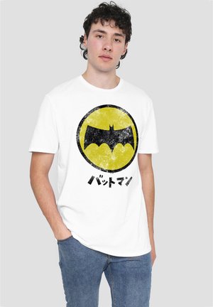 Young man with curly hair stands with hand in pocket wearing white t-shirt with black bat symbol in yellow circle and Japanese text below.