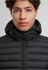 Čierna vypchaná bunda s vysokým golierom a kapucňou, s hladkou textúrou a vertikálnym prešívaním. Zapínanie na zips vpredu.