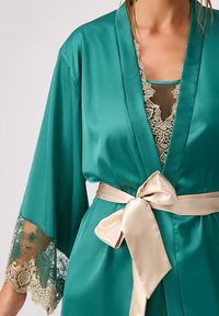Jedwabny kimono w kolorze turkusowym z koronkowymi rękawami i beżowym satynowym paskiem zawiązanym w talii. Posiada skomplikowane koronkowe detale na warstwie wewnętrznej.