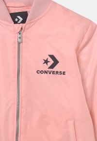 Różowa kurtka z zamkiem błyskawicznym, wyposażona w prążkowany kołnierz, czarny haftowany logo Converse z gwiazdą i chevronem na piersi oraz szczegóły w postaci bocznych kieszeni.