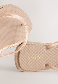 Paire de sandales beige type tongs avec des lanières pailletées et des semelles texturées, nom de la marque "NEXT" embossé en or sur la semelle intérieure.