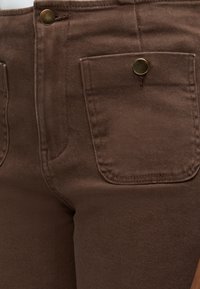 Gros plan sur un pantalon marron avec fermeture à bouton-pression à l'avant et une poche avant sécurisée par un bouton-pression sur le côté droit.