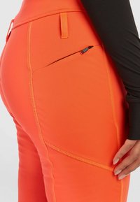 Hoogsluitende, oranje legging met een zijzak met rits, contrasterende stiksels en een gladde, rekbare stof voor comfort en bewegingsvrijheid.