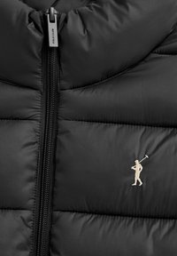 Czarna pikowana kurtka z centralnym zamkiem błyskawicznym i małą beżową haftowaną postacią trzymającą kij do polo po prawej stronie.