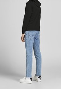 Man som bär ljusblå jeans, vita sneakers och en svart hoodie, står med ryggen mot kameran framför en enkel grå bakgrund.