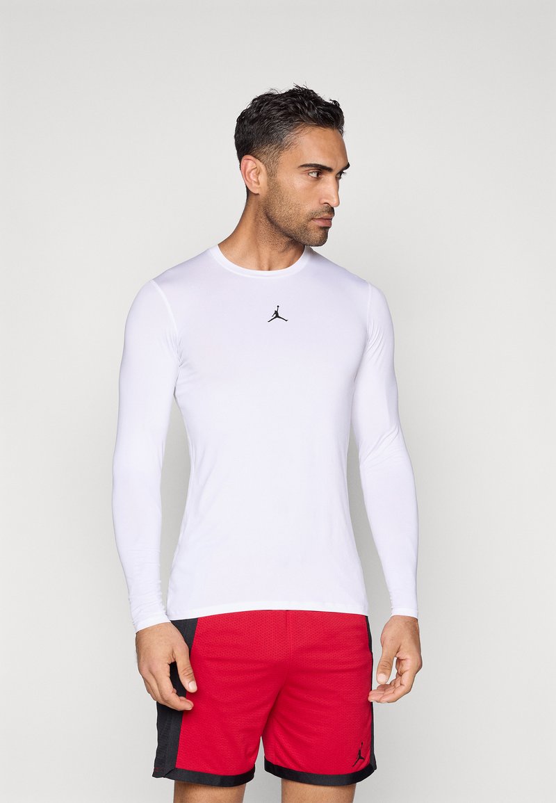 Hvit langermet treningsskjorte i glatt materiale, med en svart Jordan-logo midt på, kombinert med røde og svarte shorts.