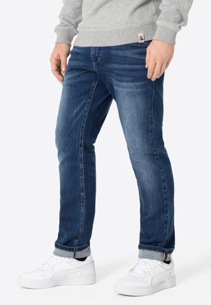 Mann trägt blaue hochgekrempelte Jeans, weiße Turnschuhe und einen grauen Sweatshirt, steht vor einem einfarbigen weißen Hintergrund.