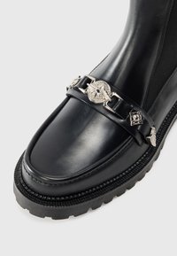 Botte en cuir noir avec une finition polie, featuring une chaîne en argent décorative près de la cheville et une semelle en caoutchouc texturé pour l'adhérence.