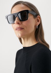 Sorte, overdimensjonerte solbriller med tykke rammer og mørke linser. Merkevarenavn "Gucci" i gull på siden. Modell iført en stripete svart topp.