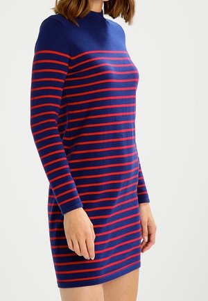 Femme portant une robe bleu marine ajustée à manches longues avec des rayures rouges horizontales, debout devant un fond clair uni.