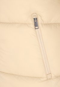 Bézs steppelt dzseki közeli kép egy függőleges cipzáras zsebbel, ezüst fém cipzárhúzóval, steppelt anyagon.