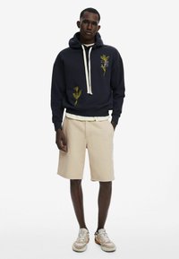 Mørkeblå pullover hoodie med cremefarvede snore og blomsterbroderi, parret med beige shorts. Model iført lyse sneakers.