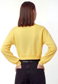 Dzeltena cropped džemperis ar garām piedurknēm, ribotu apdari un mīkstu tekstūru, kopā ar melnām biksēm ar aizmugurējām kabatām.