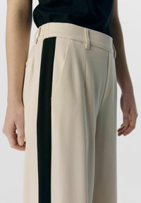 Pantaloni a gamba larga beige con una striscia laterale verde scuro, realizzati in un tessuto testurizzato e caratterizzati da pieghe frontali classiche. Accentuati da passanti per cintura.