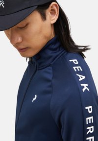 Marineblå sportsjakke med høj krave, lynlås foran og hvid "PEAK PERFORMANCE" logo vertikalt på ærmet. Glat, letvægtsstof.