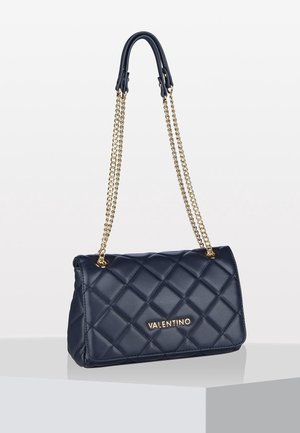Sac en cuir matelassé bleu marine de Valentino avec une bandoulière en chaîne dorée et un logo, posé sur une surface blanche avec un fond uni.