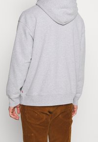 Haut à capuche en coton gris avec une poche kangourou, des poignets et un ourlet côtelés ; porté avec un pantalon en velours côtelé marron présentant des détails sur les poches arrière.