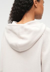 Gaišs beidžs džemperis ar kapuci, ar teksturētu adījumu, redzamām plecu šuvēm un gludu apdari uz auduma.