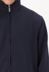 Jachetă din fleece de culoare bleumarin, cu guler înalt, închidere cu fermoar și buzunare laterale. Prezintă un logo brodat discret "LES DEUX" pe piept.
