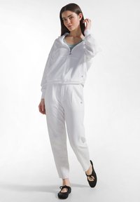 Ung kvinde iført hvid hættetrøje med lynlås og matchende bukser står med det ene ben fremme, rører ved håret og har sorte ballet flats på.