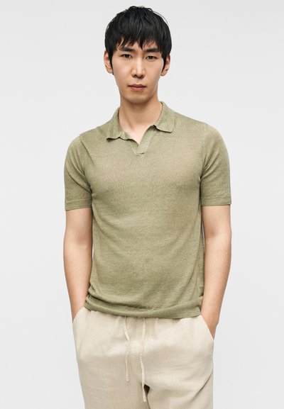Polo à manches courtes vert olive avec un col classique, un ourlet côtelé et un tissu tricoté texturé. Associé à un pantalon de couleur claire.