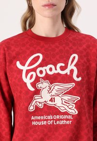 Červená mikina s texturovaným vzorem, bílým logem "Coach" a grafikou okřídleného koně s číslem "41" pod ním.
