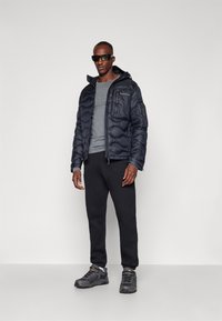 Casaco puffer azul escuro com um design acolchoado e capuz. Usado sobre uma camisa de manga longa cinza e calças de moletom pretas. Apresenta óculos de sol e sapatilhas desportivas.