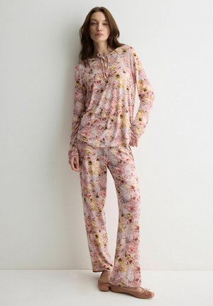 Frau trägt ein langärmeliges Top mit pinkem Blumenmuster und passende Hose, steht entspannt mit den Händen an einer weißen Wand.