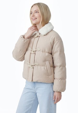 Ung kvinde smiler, iført en beige polstret jakke med lukninger med knapper og en hvid imiteret pelskant, kombineret med lyseblå jeans.