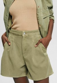 Vihreät shortsit rentoa istuvuutta, joissa on etupaneelin laskokset, sivutaskut ja nappikiinnitys. Yhdistetty vaaleanbeigeen ribbipaitaan.