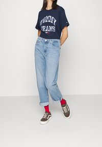 Námořnické tričko s logem "TOMMY JEANS", světle modré vysoké džíny s ohrnutými konci, červené ponožky a hnědé kostkované tenisky s bílou podešví.