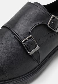 Chaussure en cuir noir avec design à double boucle, finition texturée et bout arrondi. Semelle lisse avec des accents de couture détaillés.