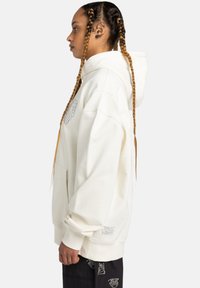 Cream hoodie with a front pocket, ribbed cuffs, and an embroidered logo. Worn with black pants featuring white graphic patterns.