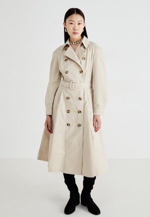 Beige trenchcoat med dobbeltbrystet front, store gullknapper, belted midje, og flagrende skjørt. Båret over svarte knehøye støvler.