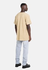 Beige Kurzarmhemd mit lockerer Passform und strukturiertem Stoff; kombiniert mit hellblauen Jeans und schwarzen Schnürschuhen; schlichtes Design.