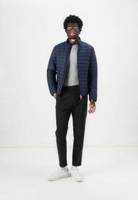 Doudoune bleu marine avec des coutures horizontales, associée à un t-shirt gris, un pantalon noir et des baskets blanches. Design décontracté, sensation légère.