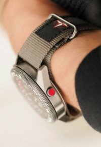 Reloj de metal plateado con una esfera redonda, botón de acento rojo y correa de tela verde con costuras negras y un parche de logo.