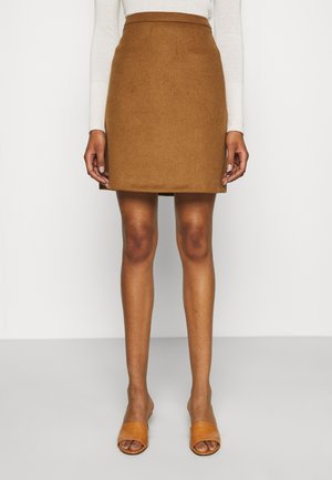 Femme portant une jupe marron longueur genou, un haut blanc à manches longues et des sandales ouvertes couleur beige, debout devant un fond blanc uni.