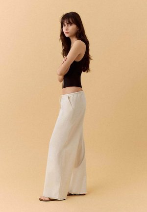 Joven mujer con cabello largo y castaño, vestida con una camiseta negra sin mangas, pantalones blancos anchos y sandalias, de pie frente a un fondo beige.