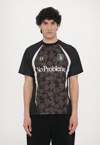 Camisola preta de manga curta com um padrão de caveira alienígena castanha, riscas laterais brancas e texto "No Problemo". O material parece leve.