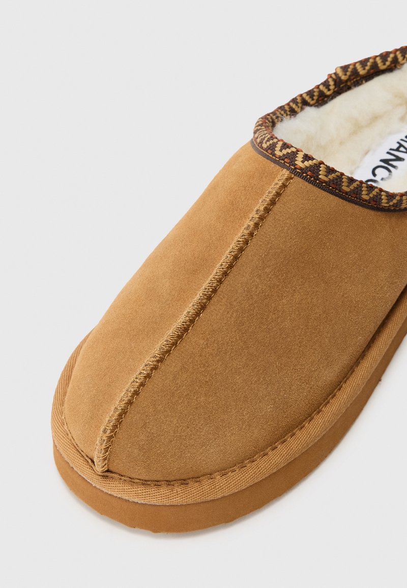 Brązowy zamszowy pantofel z teksturowaną powierzchnią, zaokrąglonym noskiem i dekoracyjnym plecionym wykończeniem wokół otworu. Wewnątrz widoczna miękka podszewka.