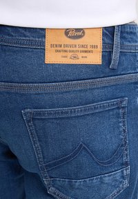 Jeans in denim blu scuro con un'etichetta in pelle sulla cintura che recita "Petrol", dotati di cuciture dettagliate sulle tasche posteriori.