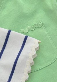 Tela verde con un bolsillo cosido y un ribete ondulado, parcialmente cubierta por tela blanca con rayas azules y un borde ondulado a juego.