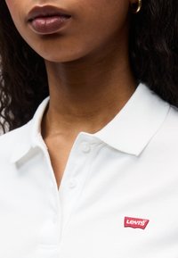 Κοντινό πλάνο γυναίκας που φοράει λευκό πόλο μπλουζάκι Levi's με γιακά και κουμπιά, δείχνοντας τα χείλη, τον λαιμό και ένα χρυσό σκουλαρίκι κρίκο.