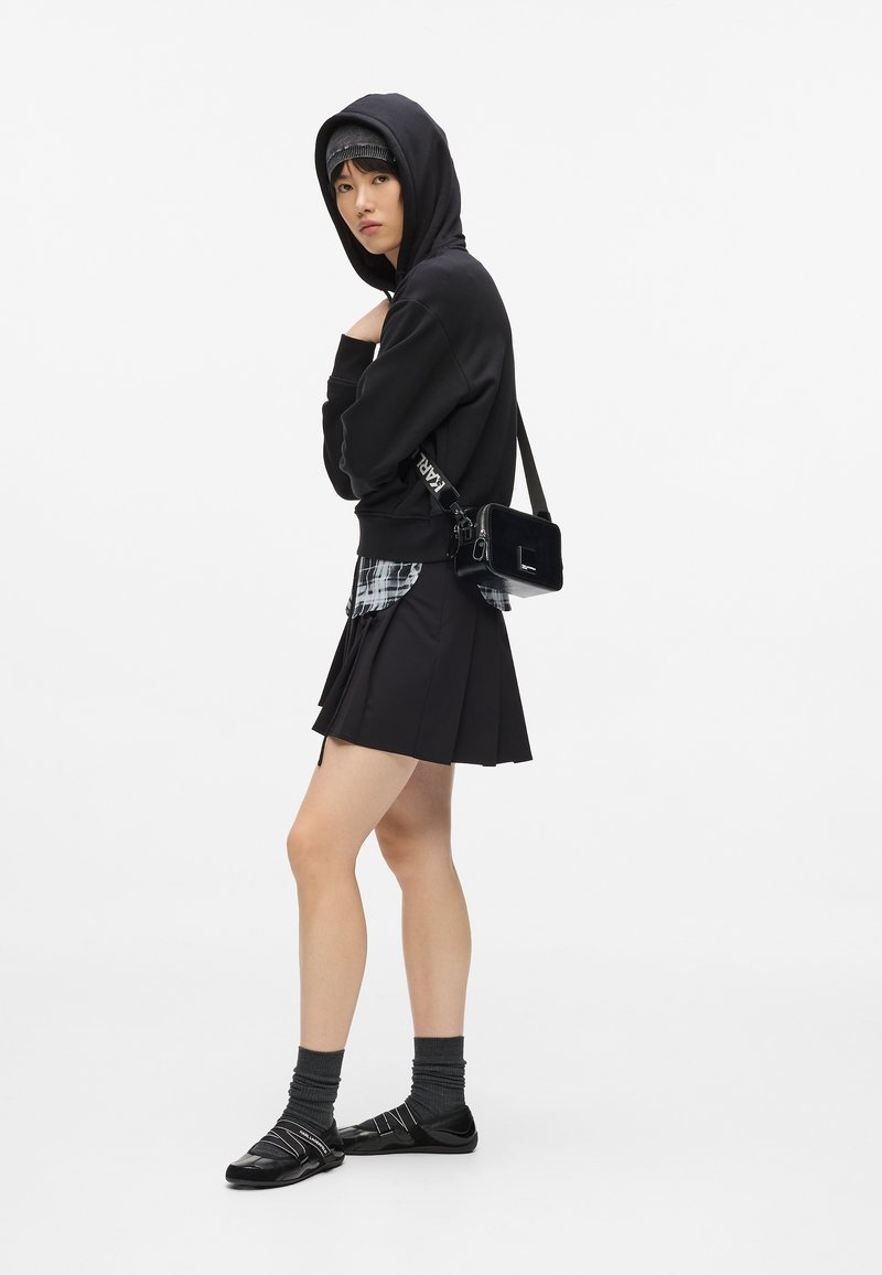 Giovane con felpa nera, camicia scozzese, gonna plissettata nera, calze grigie e scarpe nere, che porta una piccola borsa a tracolla nera.