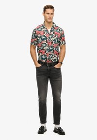 Korte mouw bloesemshirt met roze en blauwe patronen, zwarte denim jeans, zwarte schoenen en witte sokken, staand met handen in de zakken.