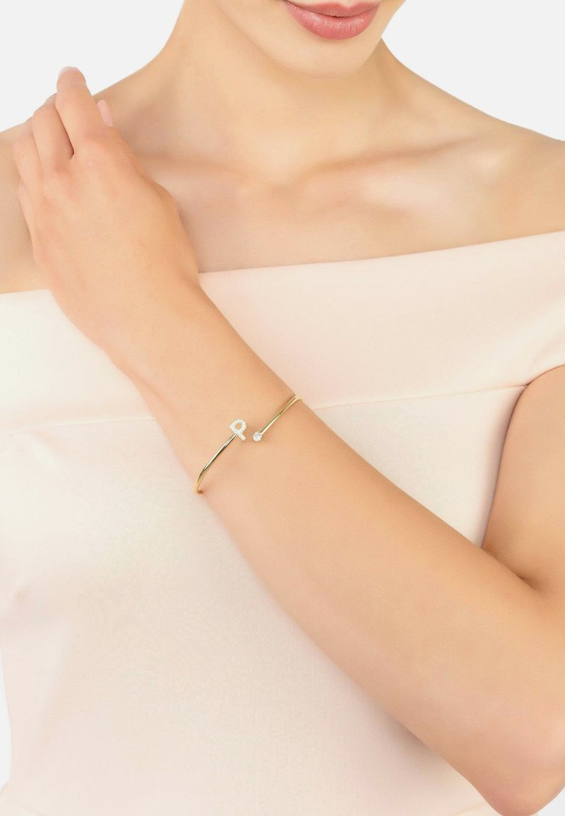 Aranyszínű karkötő sima dizájnnal, amelyen egy betű és egy kis csillogó dísz található. Minimalista és elegáns megjelenésű.