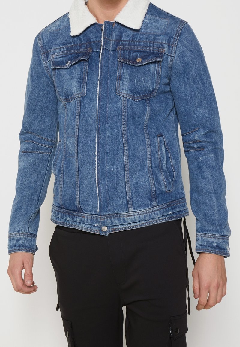 Personne portant une veste en jean bleue avec un col en shearling blanc et un pantalon noir sur fond uni.