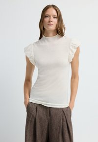 Haut crème ajusté avec des manches courtes à volants, col montant et texture lisse. Assorti à un pantalon marron plissé. Design simple et moderne.
