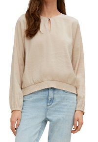 Femme portant une blouse beige à manches longues avec une petite ouverture en clé à l'avant et un ourlet élastique, associée à un jean taille haute bleu clair.